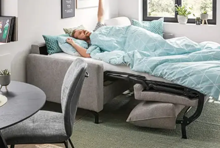Ausgezogenes Schlafsofa mit türkis gemusterter Bettwäsche, darauf liegende Person, daneben grauer Stuhl und runder Tisch, hinten Fensterbank mit Pflanzen.